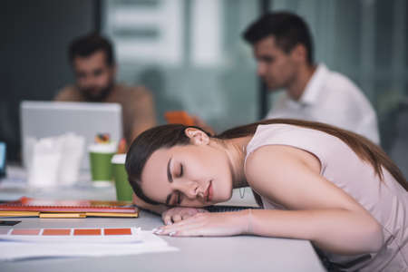 Work overload. Tired brown-haired female napping, resting her head on table, her male colleagues working behindの写真素材
