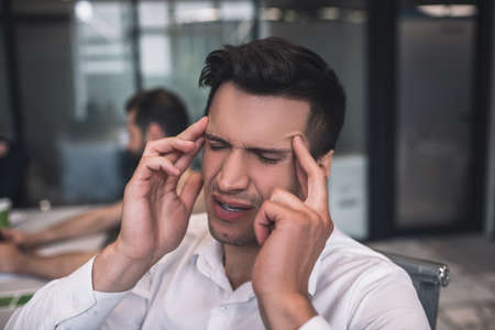 Work overload. Tired dark-haired male sitting at meeting, touching his temples, his male and female colleagues working behindの写真素材