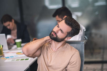 Work overload. Tired bearded male sitting at meeting, stretching his neck, his male and female colleagues working behindの写真素材