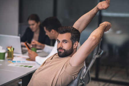 Work overload. Bearded male sitting at meeting, stretching, his male and female colleagues working behindの写真素材