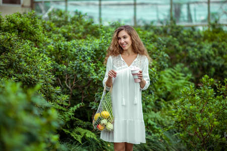 Greenhouse. Young woman in white dress with bags of vegetables and a cup in hands
