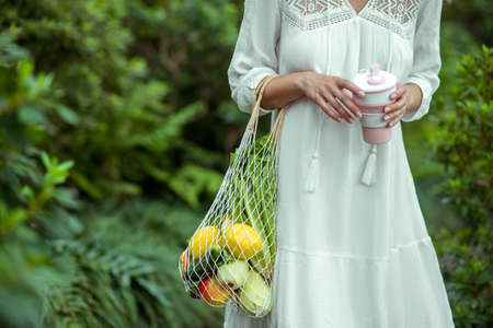 Greenhouse. Woman in white dress with bags of vegetables and no-spill cup in handsの写真素材