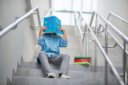School time. Schoolboy sitting on stairs, covering his face with math book