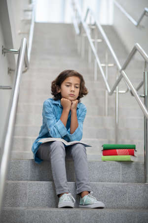 Sadness, mood. Sad boy in light blue rtsbashka and gray trousers sitting on steps of city staircase with book resting his hands on itの写真素材
