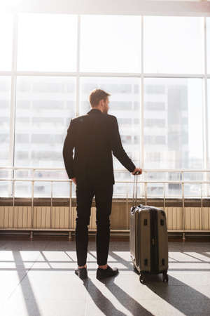 Travelling moments. A man standing with a suitcase near the huge windowの写真素材