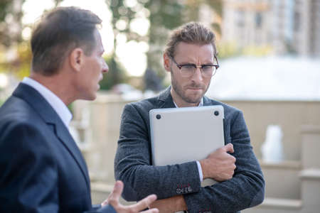Tough times. Angry businessman yelling at his younger male colleague holding laptop outside