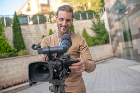 Filming process. Smiling fair-haired cameraman standing behind camera, getting ready for filming