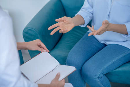 Medical appointment. Close-up of female patient hands gesticulating, female doctor sitting opposite with notebookの写真素材