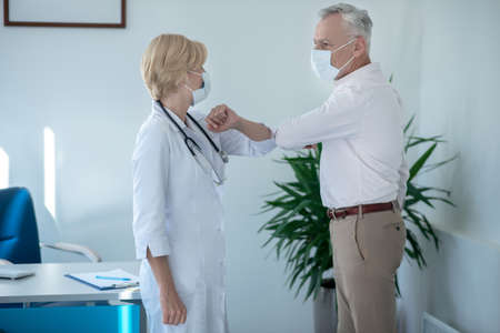 Medical advice. Gray-haired male patient touching elbows with blonde female doctor
