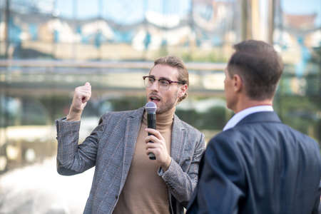 Journalistic interview. Fair-hareid male journalist speaking into microphone, interviewing mature...