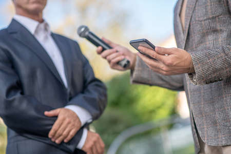 Journalistic interview. Close-up of male hands with microphone and smartphone, recording interviewの写真素材
