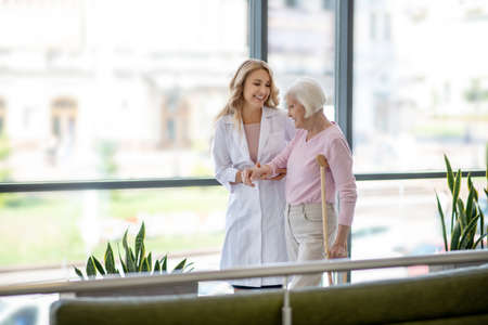 In corridor. Female doctor and an elderly woman with a crutch walking in a corridor and having a...