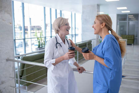 Coffee time. Two smiling female medical workers leaning on stair railing, drinking coffeeの写真素材