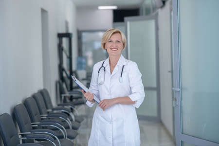 Medical profession. Smiling blonde female doctor with stethoscope standing in corridor, holding clip folderの写真素材