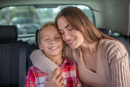 Lovely motherhood. Happy young mom embracing her daughter, sitting cheek to cheek on backseat of car