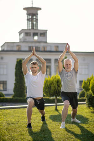 Morning workout. Young brown-haired male and mature grey-haired male stretching legs outside, holding hands above headの写真素材