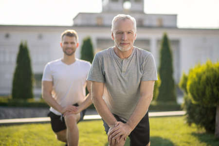Morning workout. Young brown-haired male and mature grey-haired male stretching legs outside, hands on kneeの写真素材
