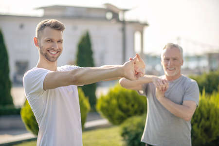 Morning workout. Young bearded male and mature grey-haired male stretching hands outside, smiling