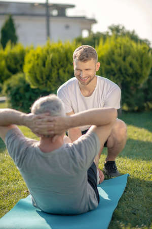 Morning workout. Smiling brown-haired male helping grey-haired male with sit upsの写真素材