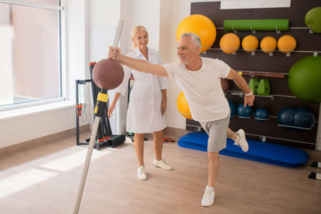 Strength. Grey-haired male patient having a workout in rehabilitation center with a female blonde instructorの写真素材