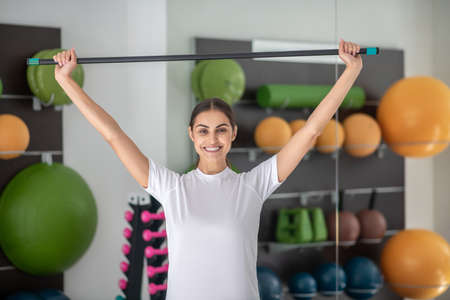 Body workout. Smiling dark-haired female stretching her shoulders with stick, hands up