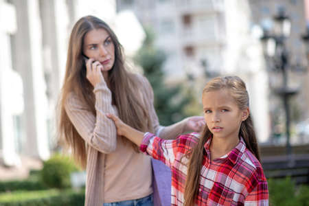 Lack of attention. Sad girl pulling her moms hand, talking on her phoneの写真素材