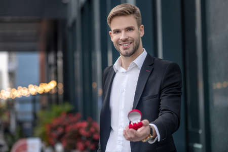 Proposal. Excited man in black suit showing a box with engagement ring