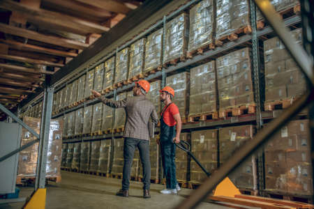 Warehouse. Manager in orange helmet talking to a warehouse worker and showing him containersの写真素材