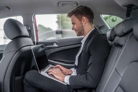 Busy schedule. Brown-haired male sitting at backseat of car, typing on laptopの写真素材