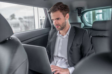 Busy schedule. Brown-haired male sitting at backseat of car, working on laptop