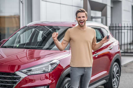 Brand-new car. Excited young male screaming with joy in front of red car