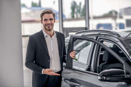 Auto dealership. Smiling brown-haired male presenting new car, holding car door in showroomの写真素材