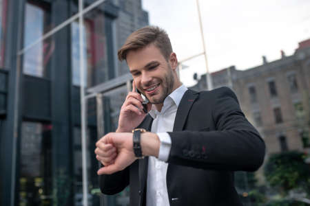 Businessman. Young man in a black suit talking on the phone and looking at his watchの写真素材