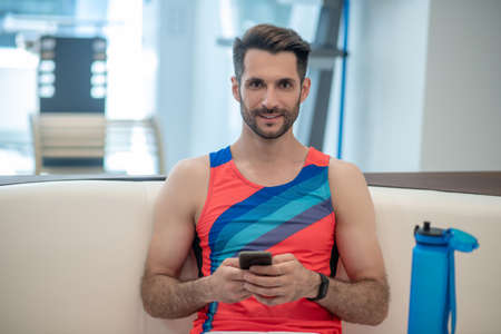 Connection. Sporty man in bright tshirt sitting with a smartphone in hands
