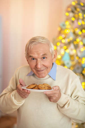 Good smell. Grey-haired man standing with a plate with pastries and smellingの写真素材
