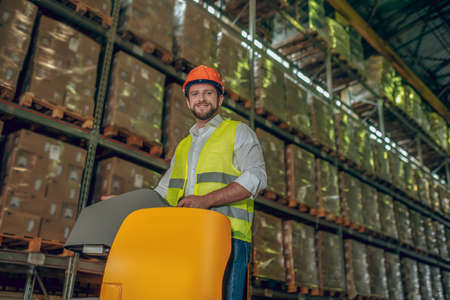 Megastore. Warehouse worker in orange helmet standing near shelves with containersの写真素材