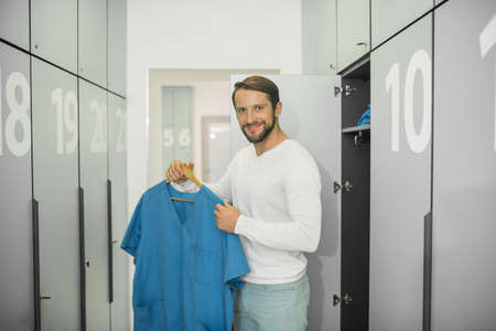 In changing room. Young male doctor standing near the locker and holding his uniformの写真素材