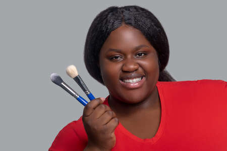 Make up. Young dark-skinned woman in red clothes holding brushes for make up.