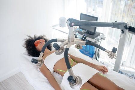 Cosmetology, laser. Young slim woman reclining on daybed receiving green laser procedure in cosmetology officeの写真素材