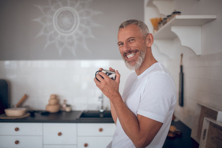 Good mood. Grey-haired tall man holding a cup and smiling nicely