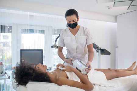 Cosmetological procedure. Young girl lying on couch with closed eyes and medic in protective mask...