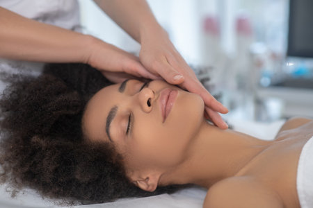 Cosmetology center. Head of young girl lying calmly on couch and hands gently massaging her faceの写真素材