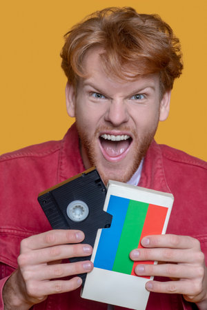 Video cassette. Red-haired bearded young man in red jacket with a video cassette