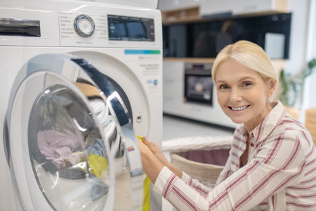 Housework. Blonde woman putting clothes into washing machine and smilingの写真素材