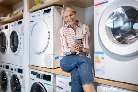 Modern housewife. Blonde woman in striped shirt sitting on a washing machine with a smartphone in handの写真素材