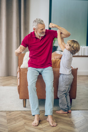 Strong man. Grey-haired man showing his muscles to his son and feeling joyfulの写真素材