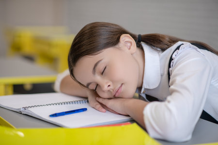 Fatigue. Cute schoolgirl with closed eyes resting head on her hands and on desk tired at schoolの写真素材