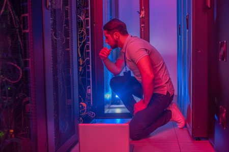 Workload. Thinking serious man in tshirt and jeans crouched near open servir rack in glowing blue...