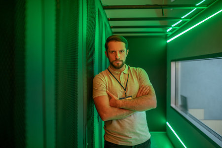 Minute of reflection. Young adult bearded man with badge standing leaning on server cabinet thinking...