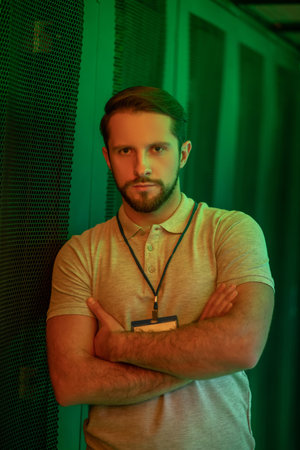 Confidence. Calm muscular young bearded man with badge standing in datacenter with arms folded on chest illuminated by green lightの写真素材